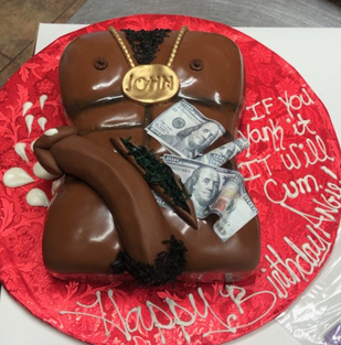 A novelty cake shaped like a muscular male torso wearing a necklace with a gold medallion. The medallion reads John. The cake has dollar bills tucked into its waistband. Text on the cake board reads, If you want it, it will cum. Happy Birthday.